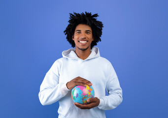 Happy black teenager protecting globe with his hands on blue studio background. Ecology, tourism,...