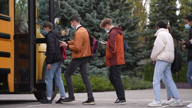 Side view of diverse high school students wearing medical masks using cellphones while standing in line to enter school bus. Yellow school bus opening doors for queue of masked secondary school pupils
