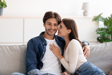 Fototapeta premium Cheerful millennial european woman kissing happy guy on cheek sitting on couch, look at camera