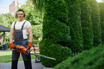 Professional male gardener wearing brown overalls, gloves and glasses standing at summer garden with electric hedge trimmer. Man enjoying seasonal work on back yard. 
