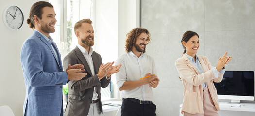Obraz na płótnie Canvas Group of happy smiling business people standing in office and clapping hands, banner background. Team of positive satisfied employees applauding to support coworker during presentation in work meeting