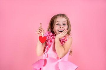 Obraz na płótnie Canvas Portrait of astonished beautiful little blue-eyed girl with long fair hair in chic pink dress stand smiling holding red paper heart on string on pink background. Valentines day, love, emotion concept.