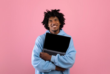 Cool black teenager in casual wear hugging laptop pc with mockup for design on empty screen over pink background