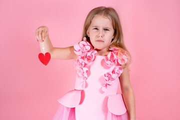 Portrait of frustrated cute little blue-eyed girl with long fair hair in chic pink dress holding red paper heart on string on pink background. Valentines day, emotion concept. Copy space, studio shot.