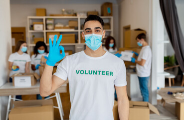 Volunteering concept. Young arab man in medical mask and gloves showing OK gesture, working in charity center