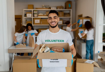 Humanitarian help. Male arab volunteer holding carton box with products, working with colleagues at charity center