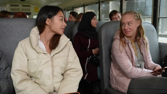Couple of multi-ethnic scoolgirls, asian and caucasian classmates enjoying conversation while riding school bus after lessons. Smiling multinational friends chatting while going home by school bus