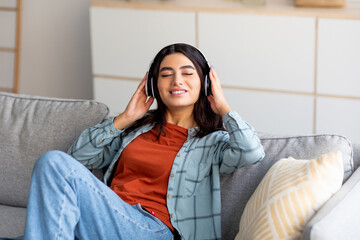 Portrait of young Arab woman in headphones relaxing on couch and listening to music with closed eyes at home