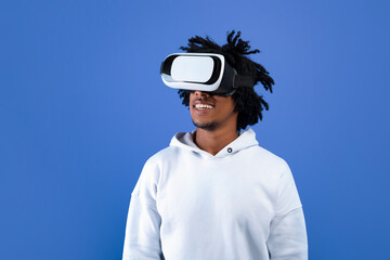 Cool African American teen guy wearing VR headset, using virtual reality for entertainment on blue studio background