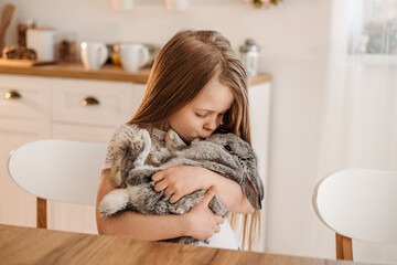 Cute little girl in a wooden kitchen at home hugging a gray rabbit in her arms
