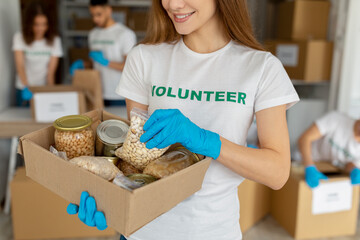 Female activist volunteer working at charity center, holding box with food donations, grains, beans...