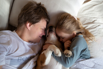 little cute girl hugging her dad and beagle dog while lying in bed