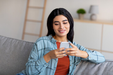 Beautiful young Arab lady using cellphone, having online meeting, sitting on sofa, browsing social network at home