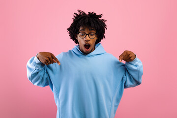 Look at this. Shocked black teen guy pointing down with two fingers on pink studio background