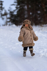 A kid in a winter fur coat, hat and felt boots in a snow-covered forest