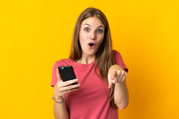 Young woman using mobile phone isolated on yellow background surprised and pointing front