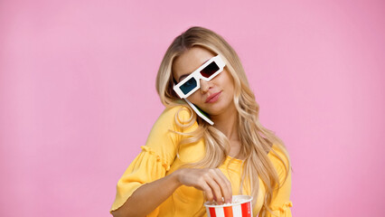 Woman in 3d glasses talking on smartphone and holding popcorn isolated on pink.