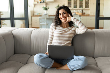 Obraz premium Cheerful young woman doing phone gesture, saying call me back, using laptop, sitting on couch at home, copy space