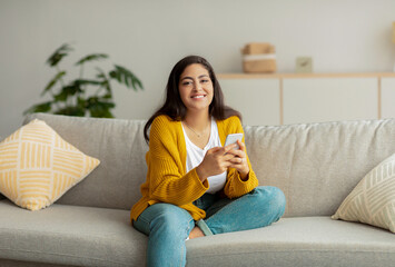 Positive middle eastern lady sitting on sofa and using smartphone, chatting with friends or lover,...