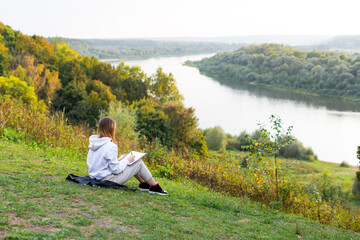 Naklejka na ściany i meble A student girl is sitting on the river bank and writing in a notebook