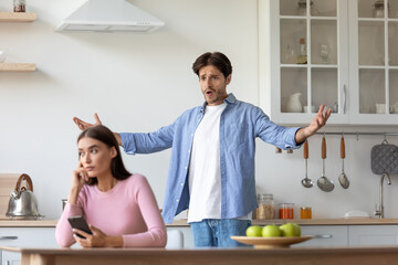 Sad young european woman with smartphone ignores offended angry screaming gesticulating husband