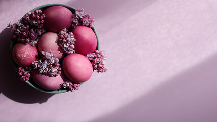 A grey bowl with bright pink Easter eggs and lilac flowers on a purple background. Easter decor. Selective focus