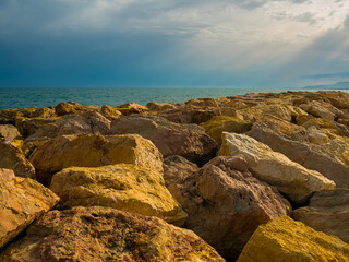 Fototapeta na wymiar Big stones of breakwater by the sea, sunlit dark clouds in background