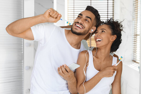 Cheerful Arab Couple With Smartphone And Earphones Listening Music While Brushing Teeth