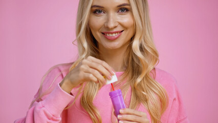 Cheerful blonde woman holding wand and soap bubbles isolated on pink.