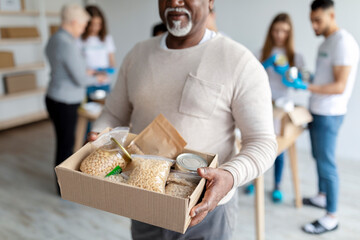 Humanitarian aid. African american elderly man holding box with donations food, volunteers packing...