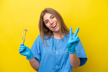 Dentist caucasian woman holding tools isolated on yellow background smiling and showing victory sign