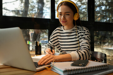 Young female student with laptop and headphones studying at table in cafe