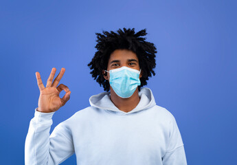 Funky black teen guy wearing face mask, gesturing OKAY, approving coronavirus protection on blue studio background