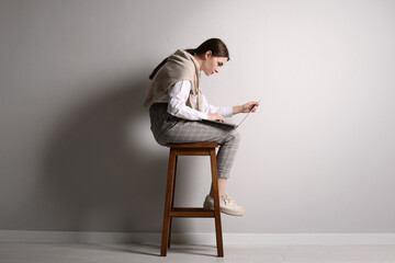 Young woman with bad posture using laptop while sitting on stool near light grey wall indoors