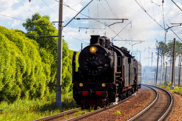 retro steam locomotive releasing smoke from the chimney, rides on the railroad, past the green trees