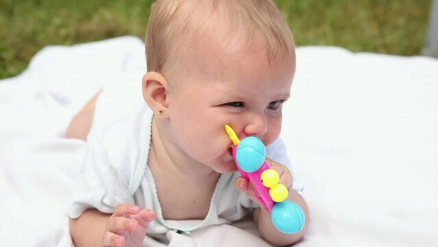 Little newborn girl lying on tummy chewing rattle on blanket, summer day outdoor. Infant having fun outdoors. Infant baby child resting playing learning to crawl. Motherhood teething, babyhood concept