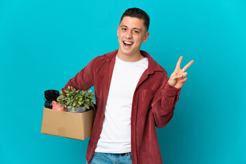 Young caucasian man making a move while picking up a box full of things isolated on blue background smiling and showing victory sign