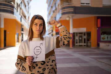Young woman holds a gender equality sign while raising her arm showing her strength in the middle of the street.