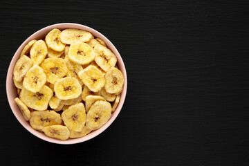 Homemade Banana Chips in a Pink Bowl on a black background, top view. Flat lay, overhead, from above. Copy space.
