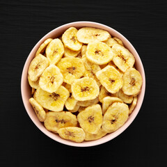 Homemade Banana Chips in a Pink Bowl on a black background, top view. Flat lay, overhead, from above.