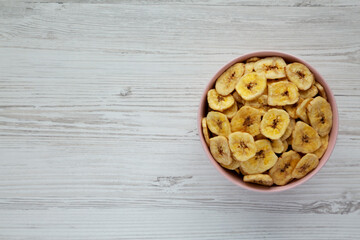 Homemade Banana Chips in a Pink Bowl on a white wooden surface, top view. Flat lay, overhead, from above. Copy space.