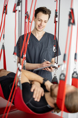 Rehabilitation specialist examining male patient before active treatment on suspension straps. Therapeutic exercises and neuromuscular activation on red cord slings