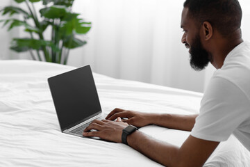 Smiling millennial african american bearded man look at laptop with blank screen, work remotely on white bed