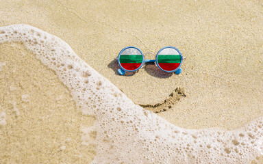Fototapeta na wymiar Sunglasses with flag of Bulgaria on a sandy beach. Nearby is a sea lightning and a painted smile. The concept of a successful vacation in the resorts of Bulgaria.