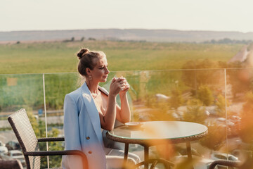 A middle-aged woman sits in a street cafe overlooking the mountains at sunset. She is dressed in a blue jacket and drinks coffee admiring the nature. Travel and vacation concept.