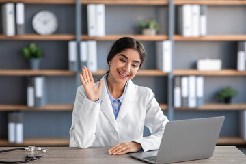 Friendly cheerful young indian female doctor in white coat works at workplace and waves hand to laptop in clinic