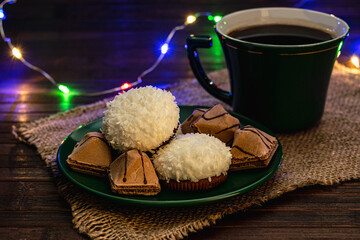 Green tea set. Coconut souffle cakes and crispy biscuits on a saucer with a cup of coffee or tea. Photo in a dark key, subdued light from the lights of a multi-colored garland.
