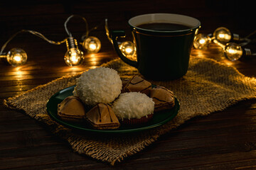 Coconut souffle cakes and crispy biscuits on a saucer with a cup of coffee or tea. Photo in a dark key, warm subdued light from the lights of a lamp garland. Green tea set.
