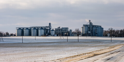 Obraz premium agro silos granary elevator in winter day in snowy field. Silos on agro-processing manufacturing plant for processing drying cleaning and storage of agricultural products, flour, cereals and grain.