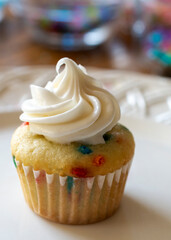 A close up view of a mini cupcake decorated with white icing.  Confetti cupcake batter on a white plate.  Wooden table and candy sprinkles blurred in background.